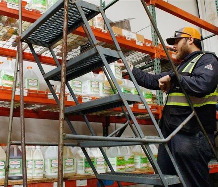 Image depicts a warehouse shelving unit assembled with cleaning products. A worker standing on a ladder inspecting them